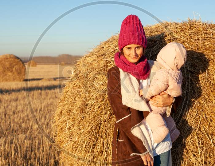Portrait Of A Beautiful Young Model In Knitted Hat  And Warm Clothes  With Little  Baby  Play, Enjoy Day, On Background Haystacks In  Sunny Autumn Day . Autumn Warm Photo. Woman Smiling And Look Away, Joyful Cheerful Mood.