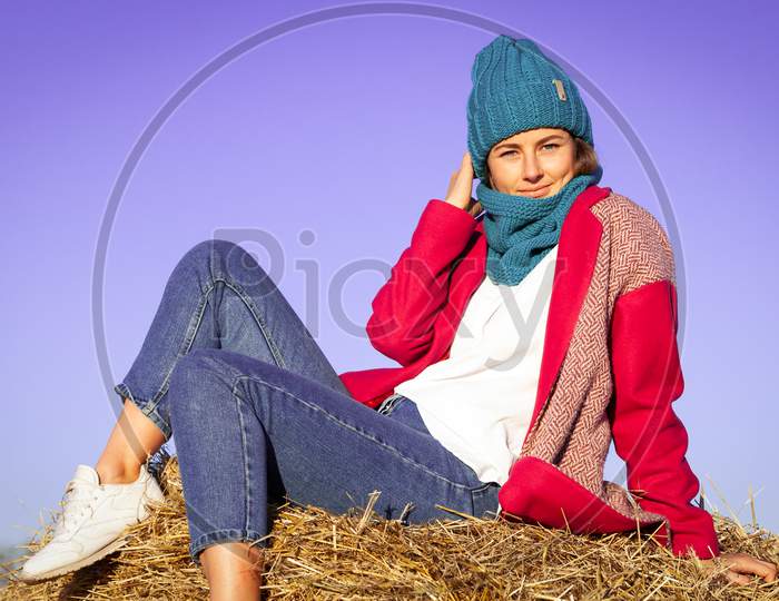 Portrait Of A Beautiful Young Model In Knitted Hat  And Warm Clothes Enjoy Day, On Background Haystacks In  Sunny Autumn Day . Autumn Warm Photo. Woman Smiling And Look Away, Joyful Cheerful Mood.