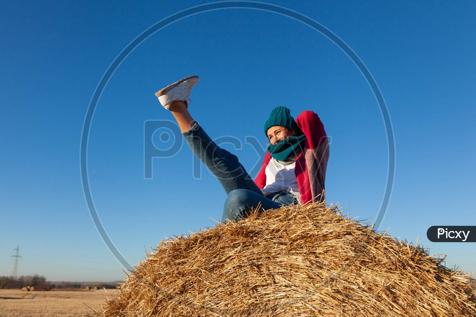 Portrait Of A Beautiful Young Model In  Blue Knitted Hat  And Warm Clothes  Have Fun,  Sitting On Haystacks In  Sunny Autumn Day . Autumn Warm Photo. Woman Smiling And Look Away, Joyful Cheerful Mood.