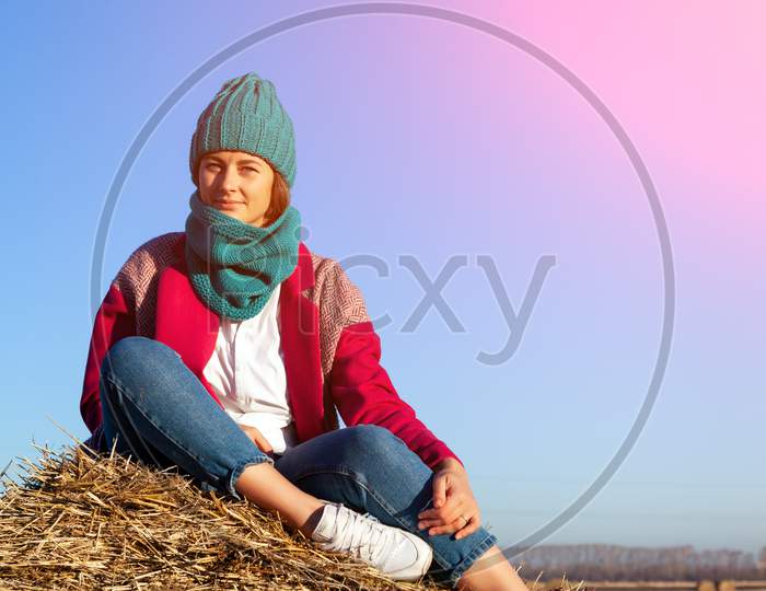 Romantic Young Woman On Blue Natural Background Outdoors. Enthusiastic Smiling Girl  In Blue Knitting Hat, Pink Coat, Shirt And Jeans Posing In A Stack Of Hay