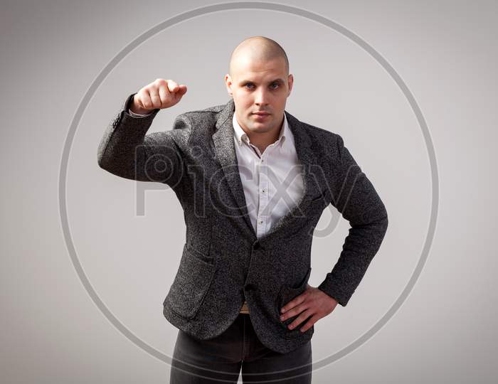Young Bald Man In White Shirt, Gray Suit Looks Confidently At Camera And Shows Index Finger In Camera On White Isolated Background