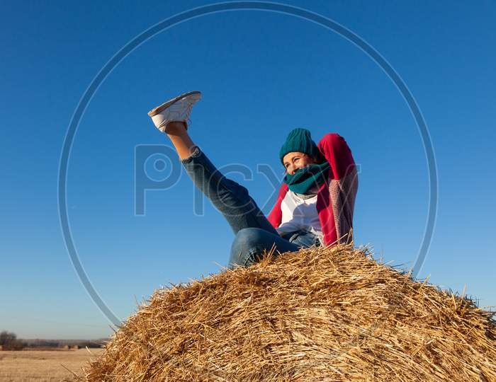 Portrait Of A Beautiful Young Model In  Blue Knitted Hat  And Warm Clothes  Have Fun,  Sitting On Haystacks In  Sunny Autumn Day . Autumn Warm Photo. Woman Smiling And Look Away, Joyful Cheerful Mood.