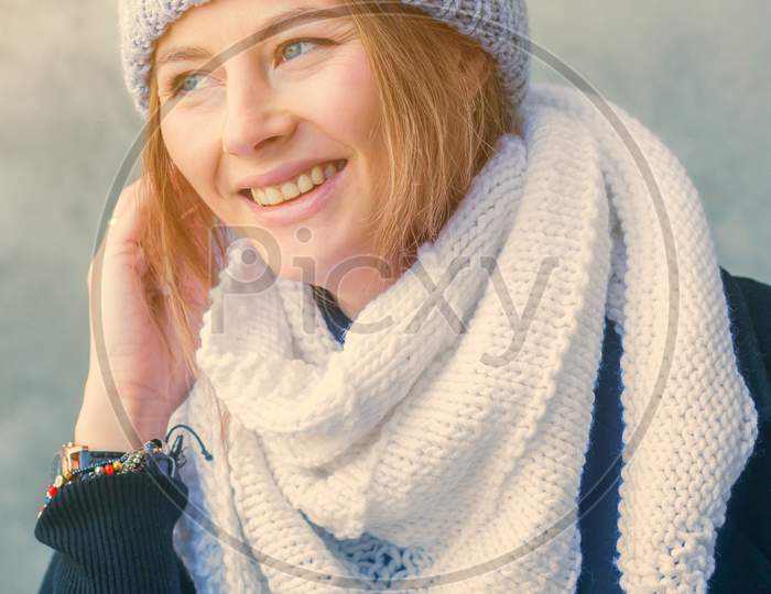A Beautiful Young Woman With Dark Hair In A Large Knitted White Bactus Scarf Made Of Natural Wool And Black Classic Сoat  Smiling And Posing On The Background Of A Stone Gray Wall