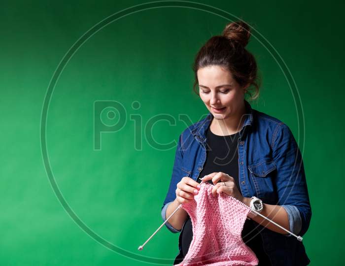 Young Woman In Denim Shirt Knits With Knitted Sweater From Natural Woolen Thread Of Pink Color On Green Isolated Background