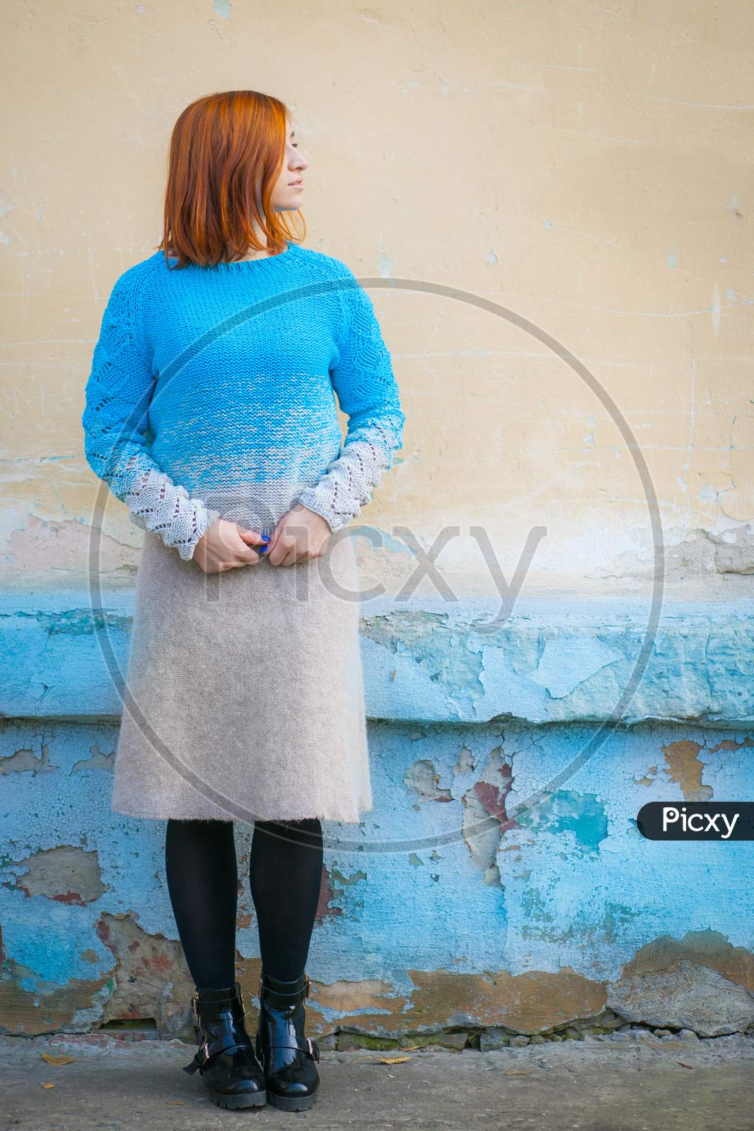 A Beautiful Young Woman With Red Hair In A Blue Sweater Made Of Natural Wool And Woolen Beige Skirt Smiling And  Posing On A City Street