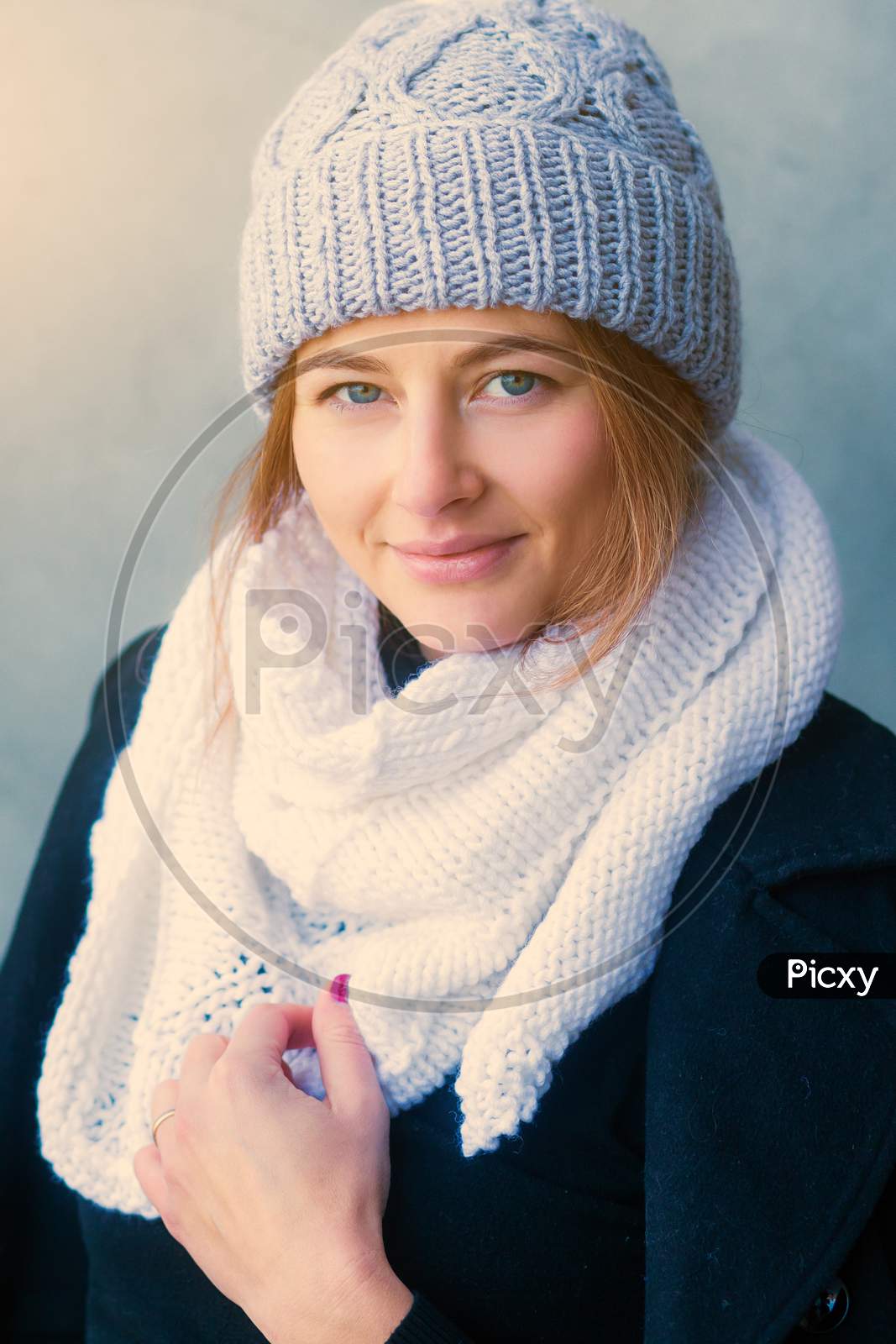 Portrait Of A Beautiful Young Woman With Dark Hair In A Large Knitted White Bactus Scarf Made Of Natural Wool And Black Classic Сoat On The Background Of A Stone Gray Wall