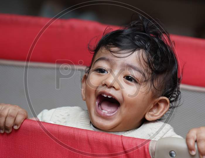 A Closeup Phot Of An Adorable Indian Toddler Baby Boy Smiling With Dimple In Cheeks And Standing Inside A Playpen.