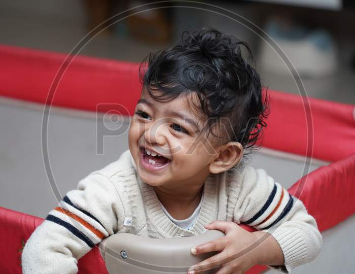 A Closeup Photo Of An Adorable Indian Toddler Baby Boy Smiling With Dimple In Cheeks And Standing Inside A Playpen