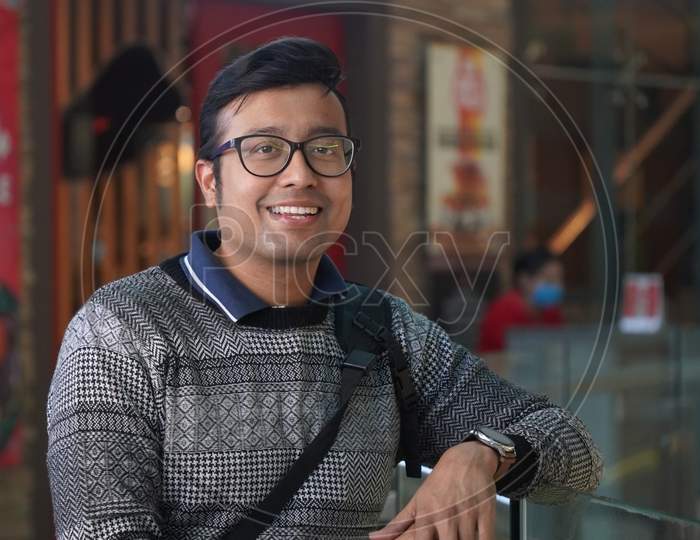 A Young Indian Male In Spectacles Looking Towards The Camera And Smiling With Blurred Background