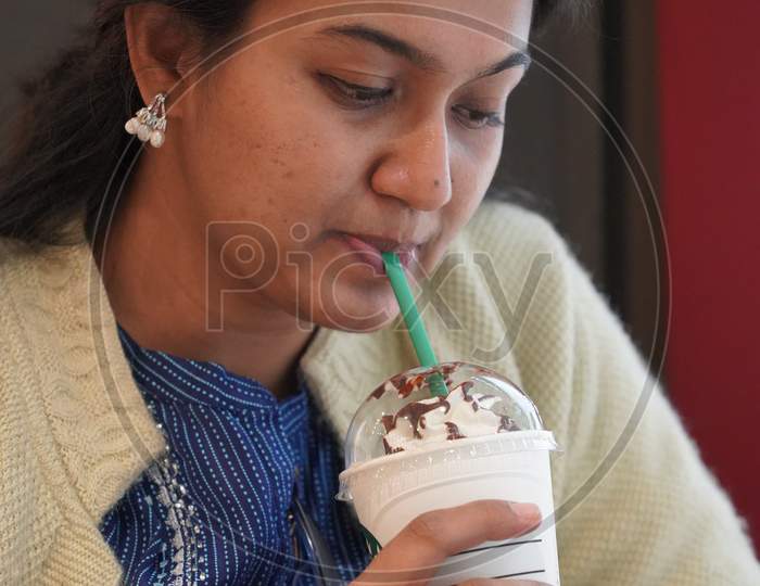 A Young Indian Lady In 30S Sipping On His Coffee Ice Cream With Straw From A Cup While Looking Down