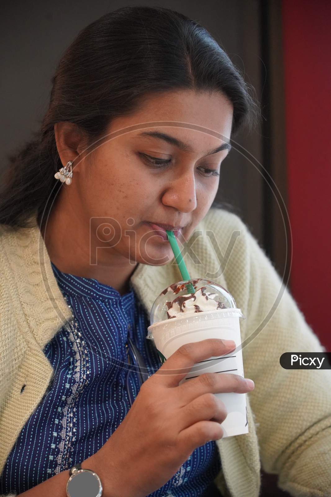 A Young Indian Lady In 30S Sipping On His Coffee Ice Cream With Straw From A Cup While Looking Down