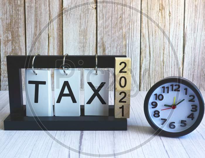 2021 number on wooden block cube and tax text on paper stand frame with black alarm clock on wooden background. Tax time concept