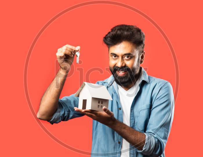 Indian Asian Bearded Young Man Holding Paper House Model With Keys And Piggy Bank