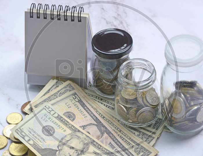 Glass jar with multicurrency coins, US dollar bills and white paper stand with space for text. Saving concept
