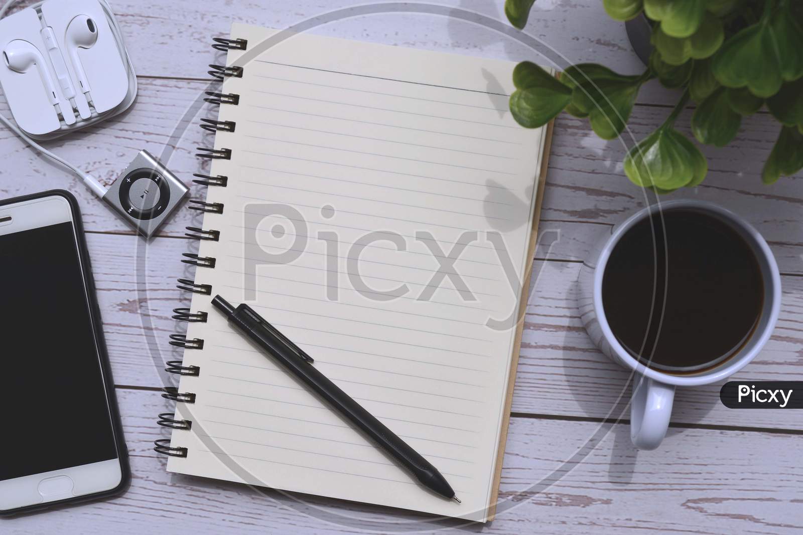 Directly above view of wooden desk with cup of coffee, smart phone, notepad, music player, pen and green leaf background