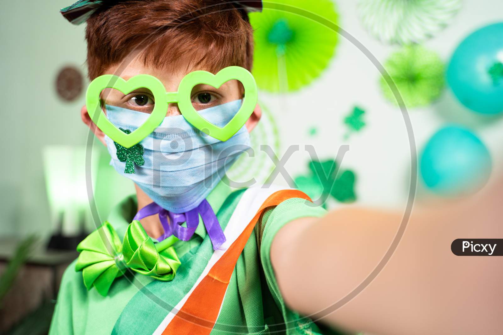 Selective Focus On Mask, Young Kid With Medical Face Mask Taking Selfie During Saint Patricks Day Celebration At Home On Decorated Background During Coronavirus Covid-19 Pandemic At Home.