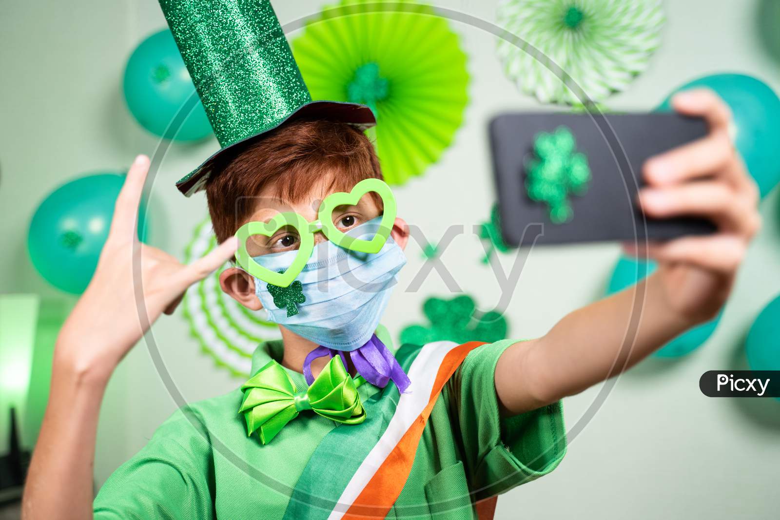 Young Kid With Medical Face Mask Taking Selfie During Saint Patricks Day Celebration At Home On Decorated Background During Coronavirus Covid-19 Pandemic At Home.