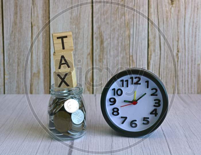 Tax text on wooden block cube on top of glass jar with multicurrency coins and alarm clock. Tax time concept