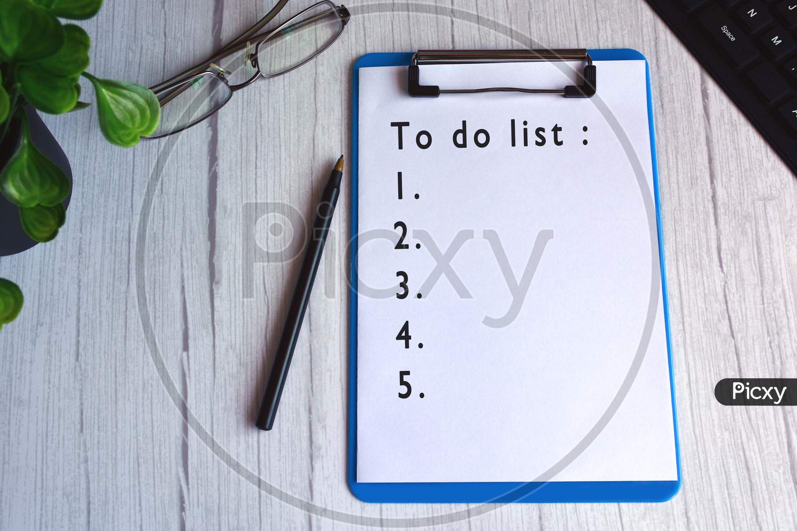 To do list text on blue clip board with glasses, pen, plant and keyboard on wooden desk.