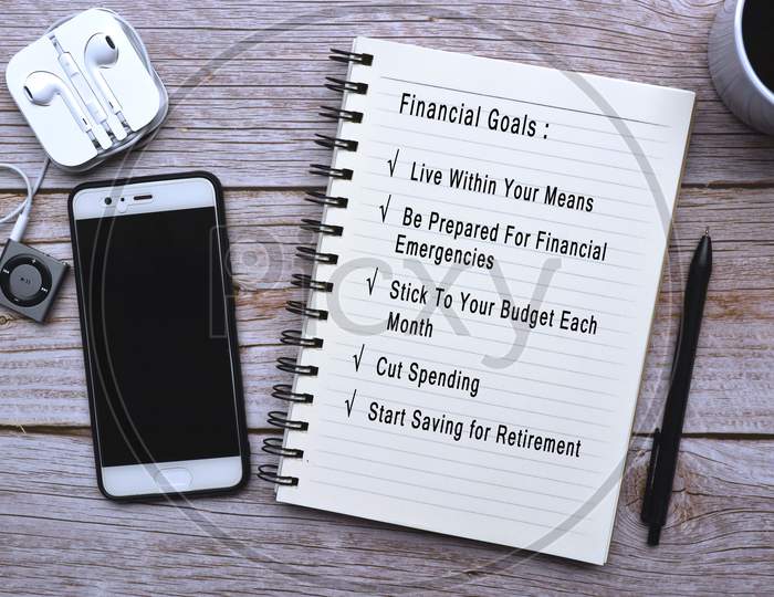 Text on notepad with cup of coffee, smart phone, music player and pen on wooden desk - Financial goals list