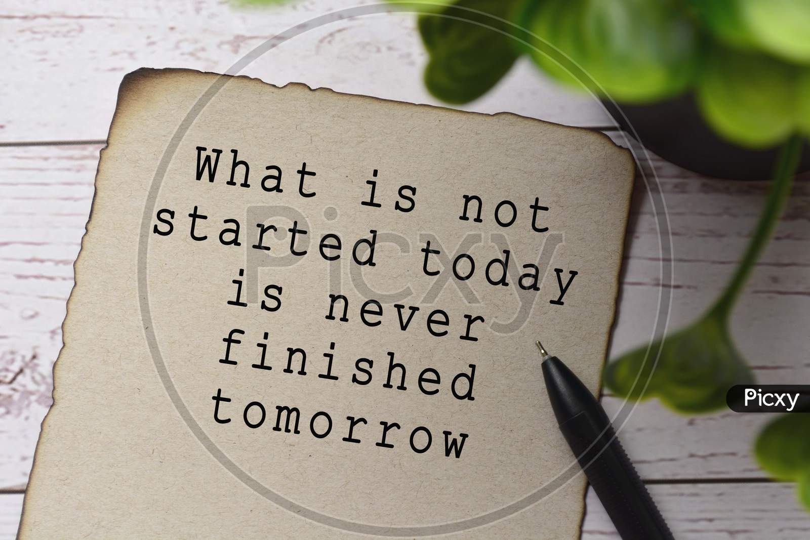 Motivational and inspirational quote on burnt edge brown paper with blurred green plant on wooden desk - What is not started today is never finished tomorrow