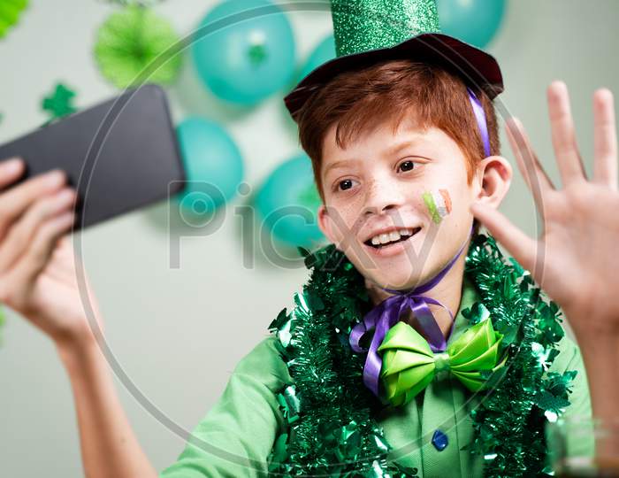 Young Kid In Green Attire Celebrating Saint Patricks Day By Making Video Call On Mobile Phone On Decorated Background During Coronavirus Covid-19 Pandemic.