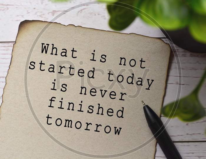 Motivational and inspirational quote on burnt edge brown paper with blurred green plant on wooden desk - What is not started today is never finished tomorrow