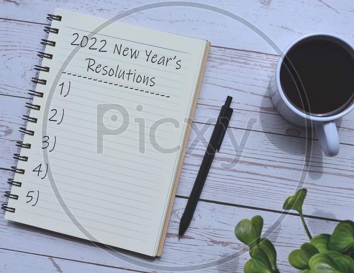 2022 New Year's Resolutions list on notepad with a cup of coffee, pen and green leaf background on wooden desk