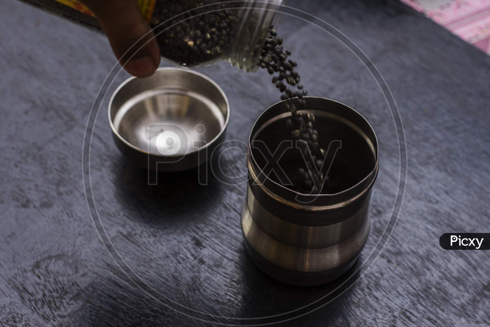 A Female Hand Pouring Black Lintel Or Kali Dal From One Steel Jar To Another Jar.