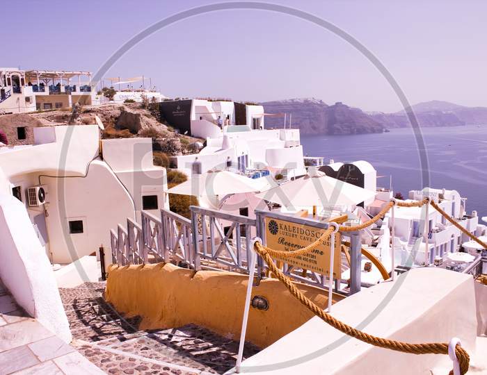 Santorini, Greece - September 11, 2017: Narrow Streets, Cyclades Architecture Hotels Houses And Cafes Over The Caldera In Oia Santorini Greek Islands Against Mediterranean Sea View With Mountains