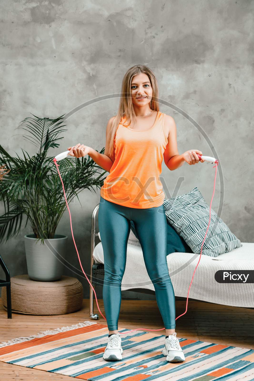 Young Woman Goes In For Sports At Home, Training Online. The Athlete Posing With Rope  , Smiles,  On Background Bed, Vase, Carpet