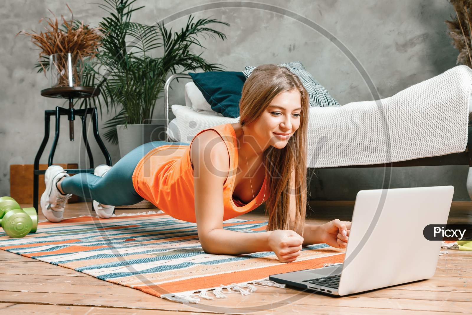 The Young Woman Goes In For Sports At Home. Cheerful Sportsman With Blond  Hair Holds The Plank And Watching Movie On Laptop  In The Bedroom
