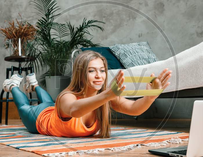 The Young Woman Goes In For Sports At Home. Cheerful Sportsman With Blond  Hair Holds The Plank With Sport Fitness Rubber Bands And Watching Movie On Laptop  In The Bedroom