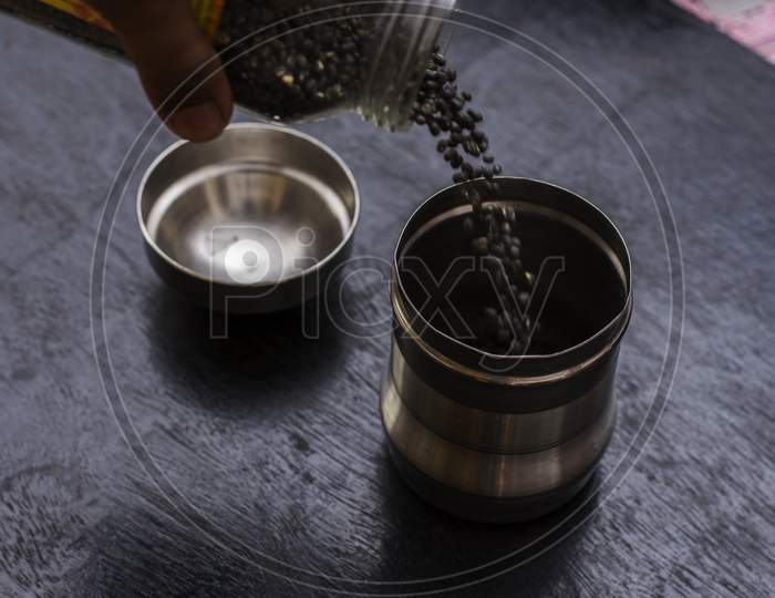 A Female Hand Pouring Black Lintel Or Kali Dal From One Steel Jar To Another Jar.