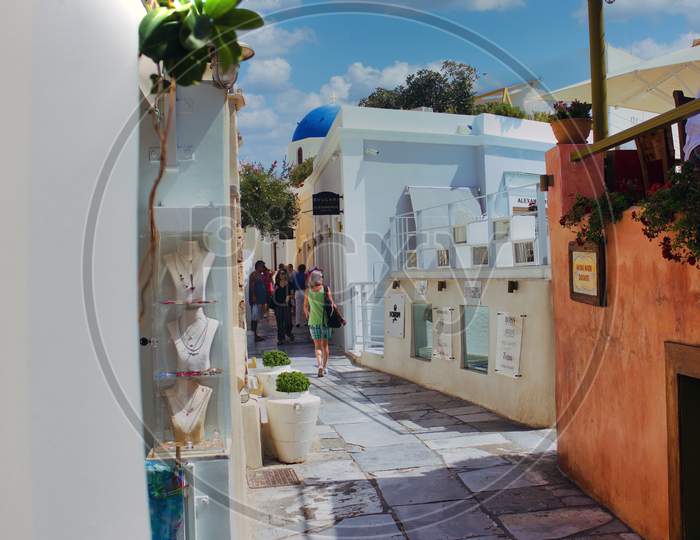 Santorini, Greece - September 11, 2017: A Narrow Commercial Street With Jewelery Shop On The Left, Tourist Walking In The Middle And People Sitting In A Restaurant On Right Against Blue Sky