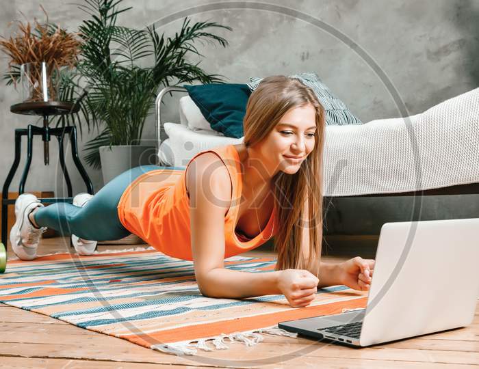 The Young Woman Goes In For Sports At Home. Cheerful Sportsman With Blond  Hair Holds The Plank And Watching Movie On Laptop  In The Bedroom