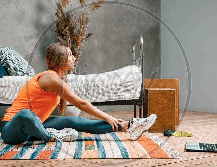 A Young Woman Goes In For Sports At Home, Online Workout . The Athlete  Stretching , Meditating, Sitting On A Twine  In The Bedroom, In The Background There Is A Bed, A Vase, A Carpet.