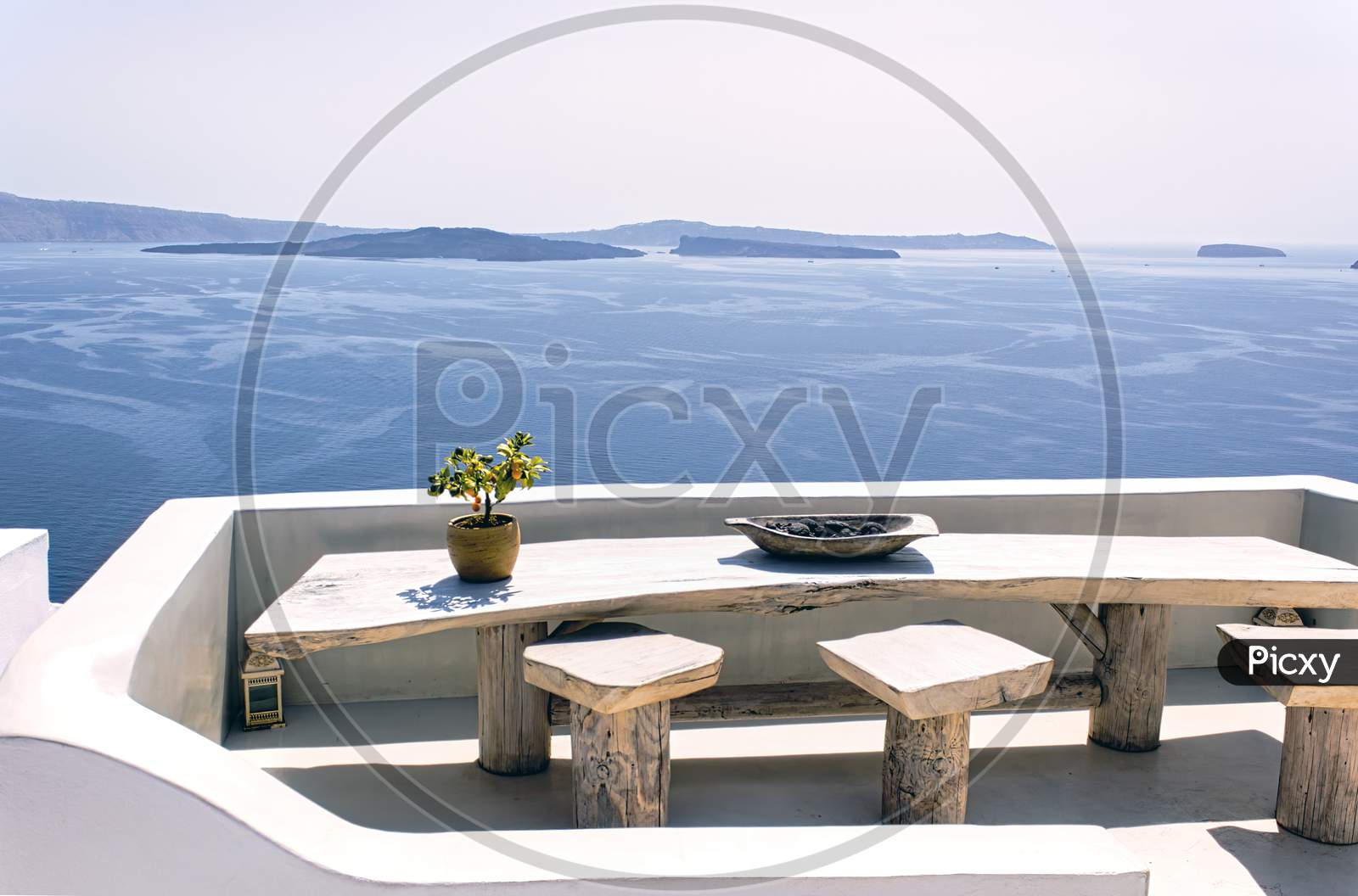 Santorini, Greece: A Pot With Flower Or Plant And A Plate On A Wooden Table With Wooden Chair Against Beautiful Sea Ocean Background With Mountains