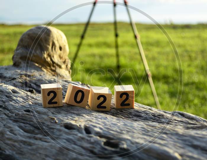 2022 Wooden Cube Block On Old Tree Stump With Blurred Background Of Green Grass Ocean And Tripod Stand
