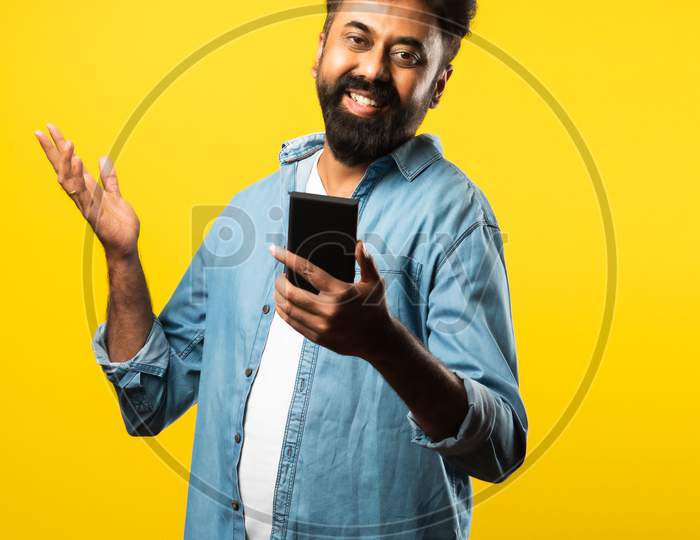 Happy Indian Bearded Young Man Using Smartphone While Standing Against Yellow Background
