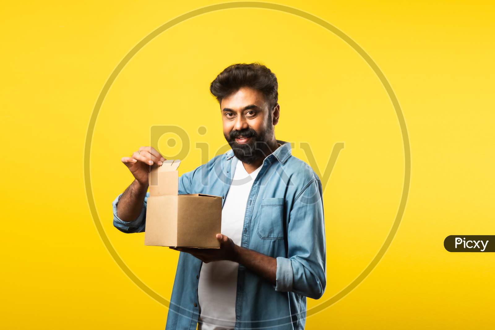 Indian Handsome Bearded Young Man Opening Box With Parcel While Standing Against Yellow Background