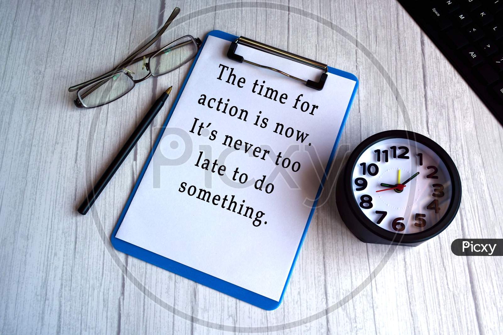 Motivational And Inspirational Quote On Blue Clip Board With Alarm Clock, Glasses, Pen And Keyboard On Wooden Desk