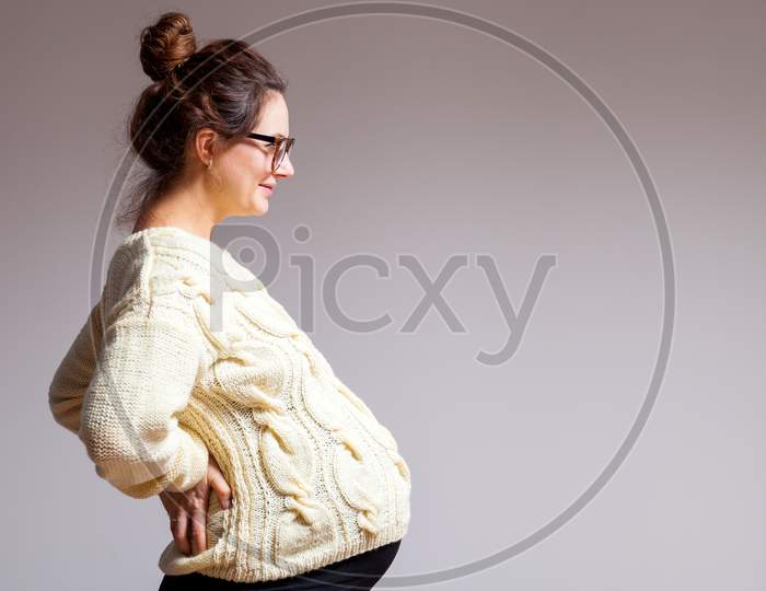 Young Dark-Haired Beautiful Woman In Late Pregnancy With Glasses And Knitted Milk-Colored Sweater Smiling And Posing On White Isolated Background