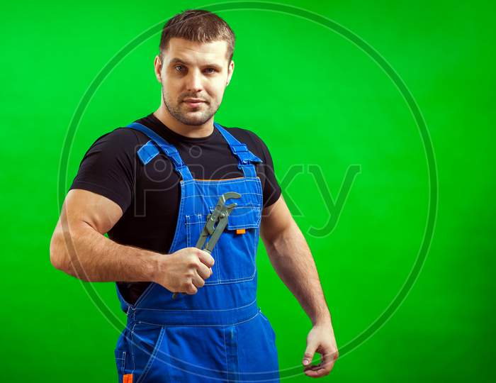 A Dark-Haired Male Construction Worker In A Black T-Shirt And Blue Construction Overall Holds A New Gray Tool  Straight Pipe Wrench On A Green Isolated Background