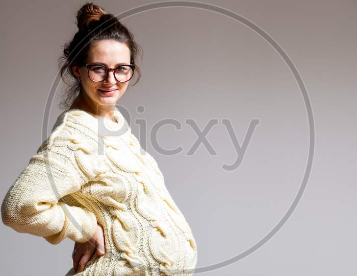 Young Dark-Haired Beautiful Woman In Late Pregnancy With Glasses And Knitted Milk-Dyed Sweater Is Holding Hands Behind Her Back On A White Isolated Background