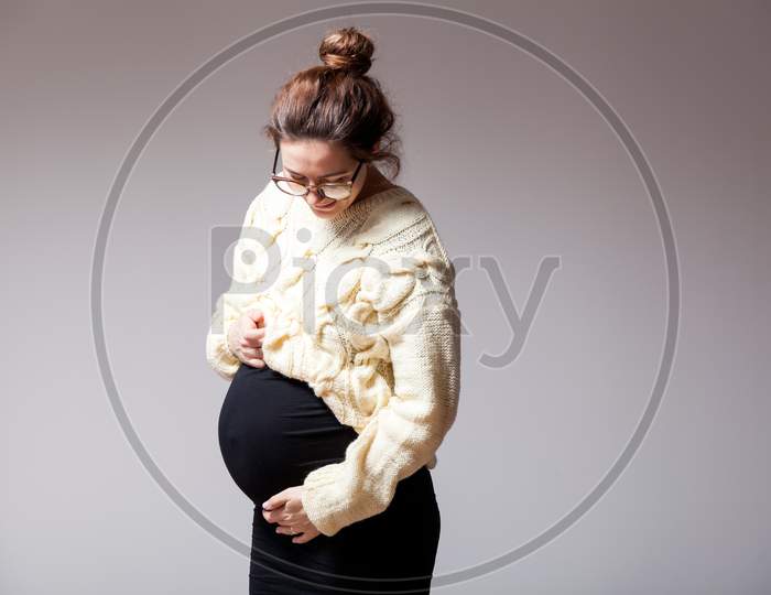 Young Dark-Haired Beautiful Woman In Late Pregnancy With Glasses And Knitted Milk-Color Sweater Posing And Holding Her Stomach On A White Isolated Background