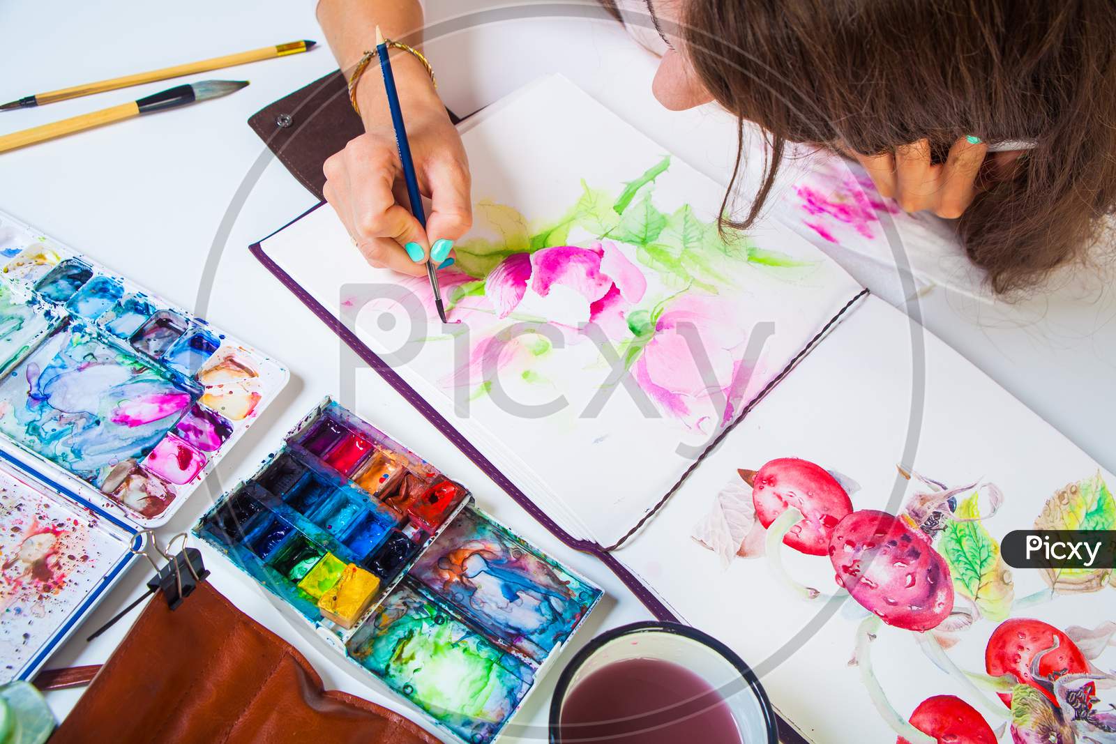 A Close-Up Artist Paints And Paints In The Album For Drawing A Thin Wooden Brush With Pink Watercolor Colors, On The Table Is A Leather Case With Brushes, Watercolor Palette,  Brushes, Cup Of Water