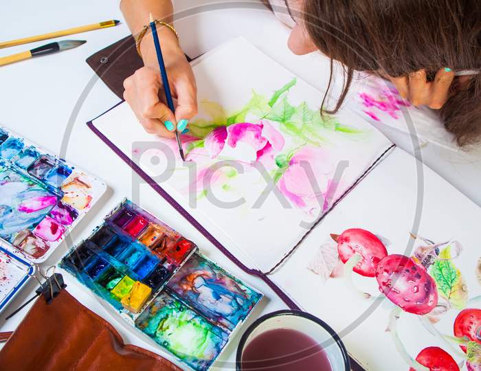 A Close-Up Artist Paints And Paints In The Album For Drawing A Thin Wooden Brush With Pink Watercolor Colors, On The Table Is A Leather Case With Brushes, Watercolor Palette,  Brushes, Cup Of Water