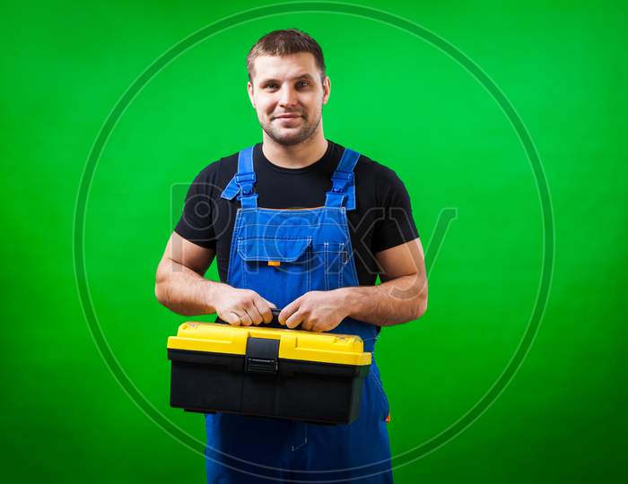 A Strong Man  Carpenter In A Black T-Shirt And Blue Construction Jumpsuit Smile And Holds  A Box With Construction Tools On His Shoulder On A Green Isolated Background