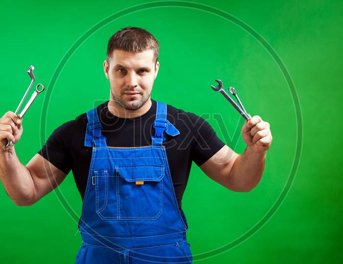 A Dark-Haired Male Construction Worker In A Black T-Shirt And Blue Construction Overall Holds In His Hands A New Tool  Professional Combination Wrench On A Green Isolated Background